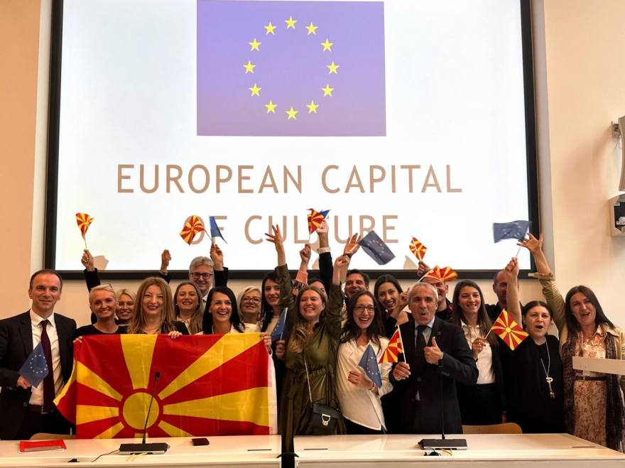 a celebratory scene with a large group of people gathered under a banner reading "EUROPEAN CAPITAL OF CULTURE." Above them is the flag of the European Union - a circle of twelve golden stars on a blue background. The people are jubilant, some holding the Macedonian flag, while others hold small EU flags. Their expressions are filled with joy and pride, clearly in a moment of celebration. This is indicative of an event celebrating Skopje's recognition as the European Capital of Culture for 2028.